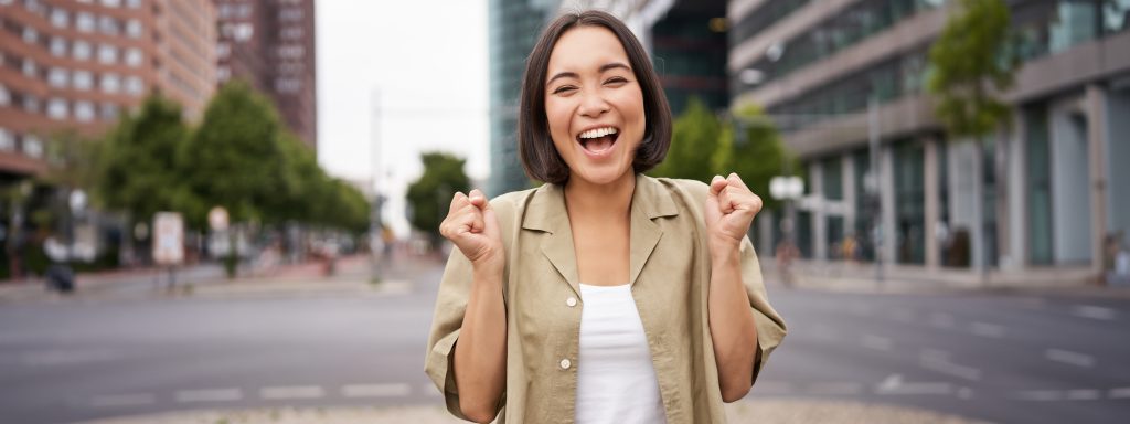 happy asian girl triumphing on streets of city, dancing from happiness, celebrating victory, posing outdoors